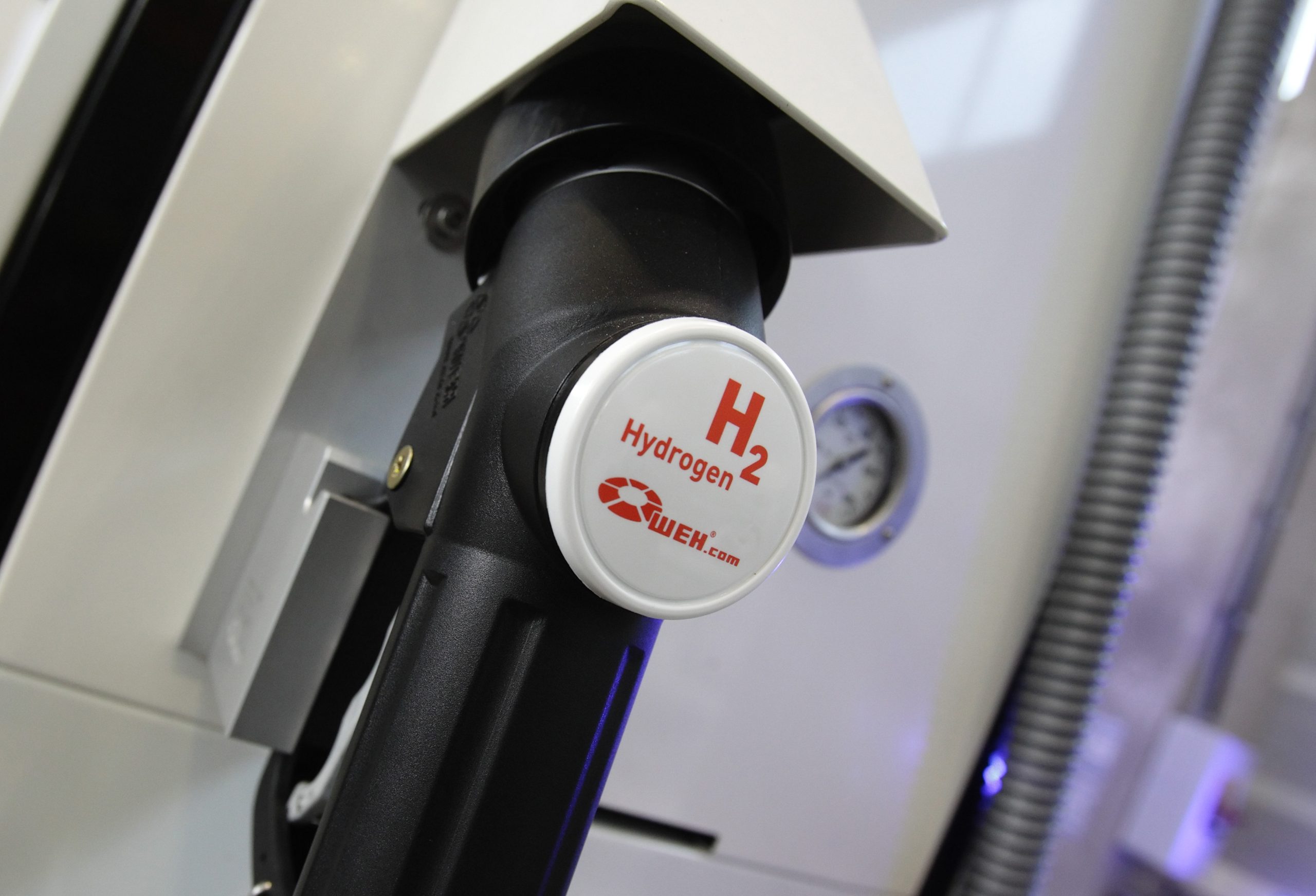 Uma bomba de combustível de hidrogênio preta, com escritos em vermelho "H2" e "Hydrogen". Ao fundo, um relógio de pressão