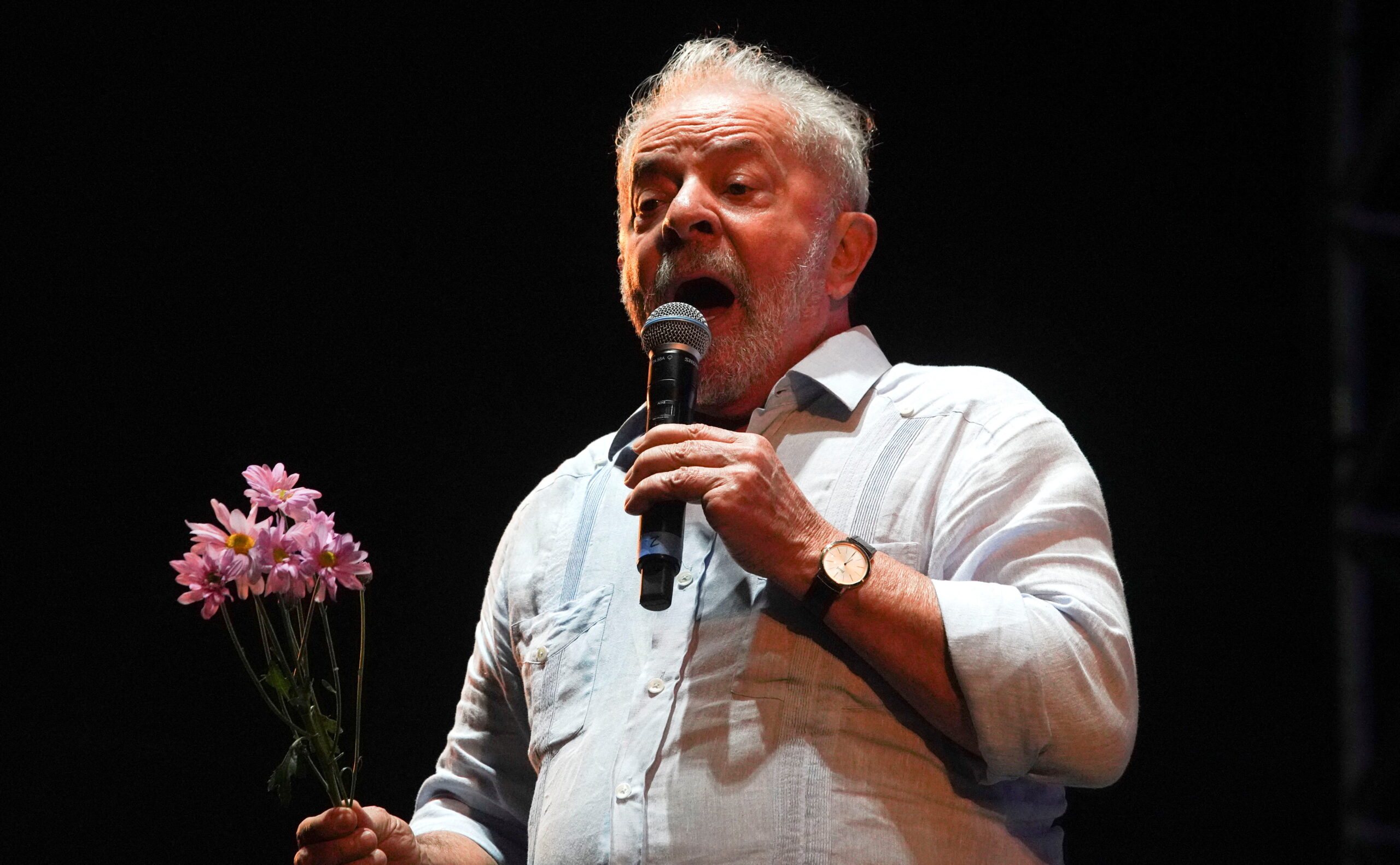 O ex-presidente Lula em evento dos 100 anos do PCdoB, no Rio de Janeiro, no dia 26 de março de 2022. Ele usa camisa branca com as mangas arregaçadas e segura um ramo de flores do campo cor de rosa na mão esquerda, e um microfone na direita
