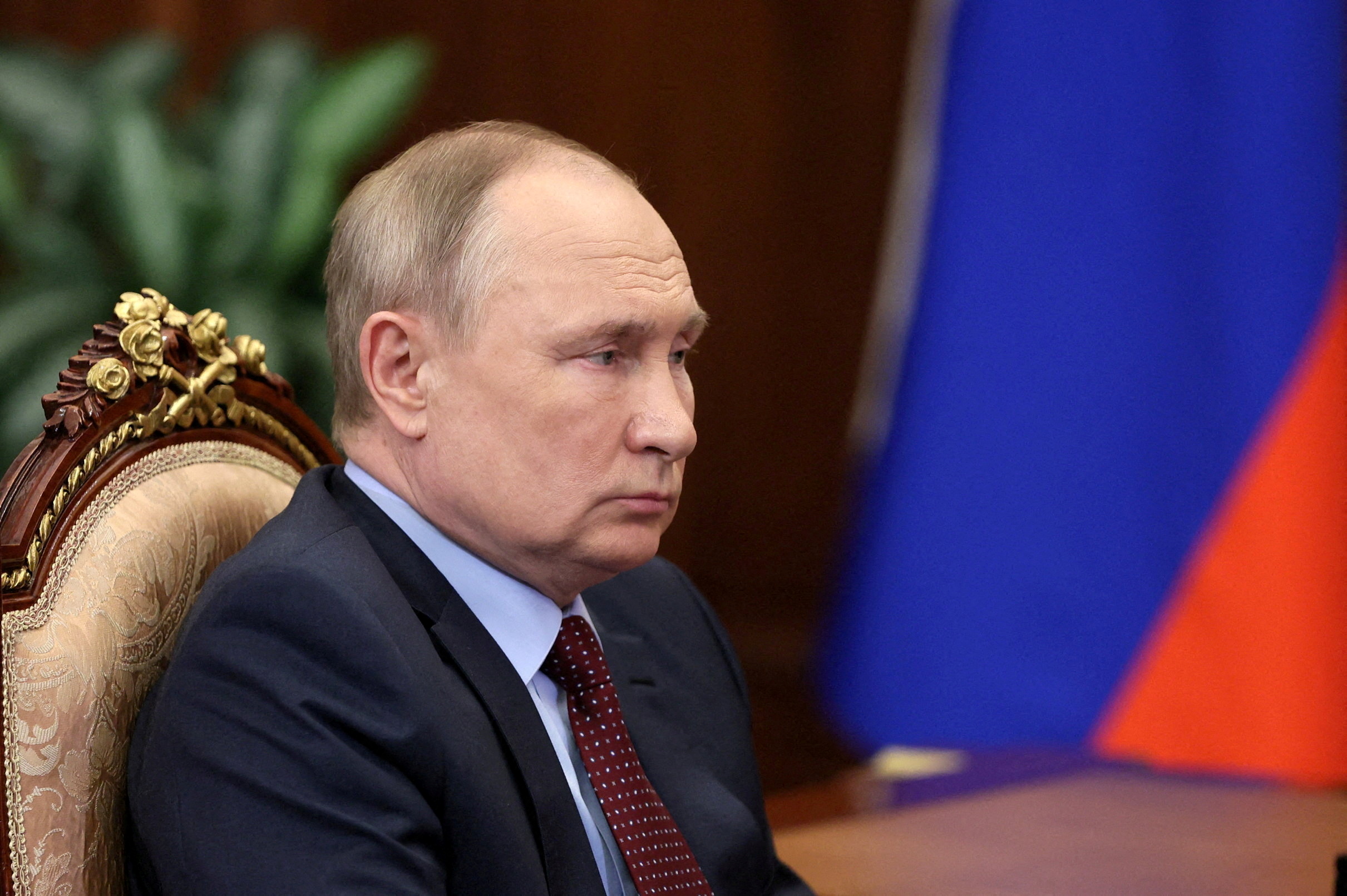 Imagem mostra o presidente russo, Vladimir Putin, em primeiro plano, de perfil, em reunião no Kremlin. Ele está sentado em uma cadeira com adornos dourados e com a bandeira russa ao fundo