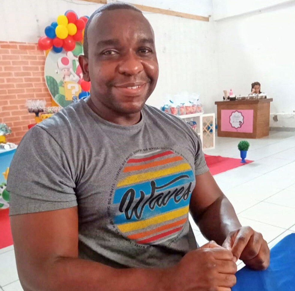 Homem negro, careca, com camiseta cinza, sorri para a foto. Ele está sentado à mesa, em um espaço que parece ser o de uma festa infantil.