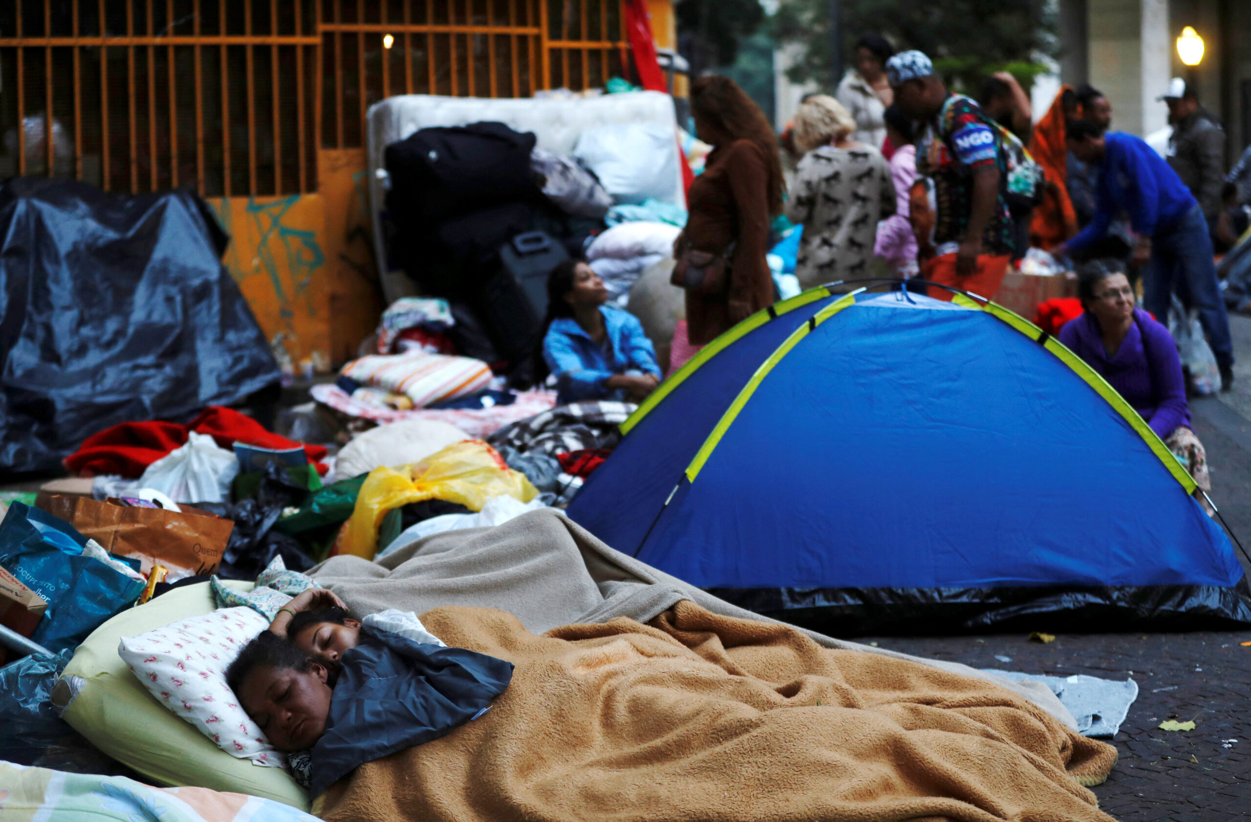 Imagem mostra pessoas dormindo em colchões na calçada, perto de barracas montadas para abrigar famílias