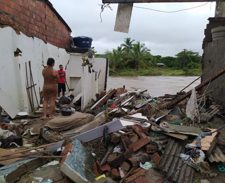 Casal dentro de casa sem telhado. Dentro da área da casa, há destroços de telhados, colhões e madeiras quebradas. Ao fundo, um rio com água e nível alto.