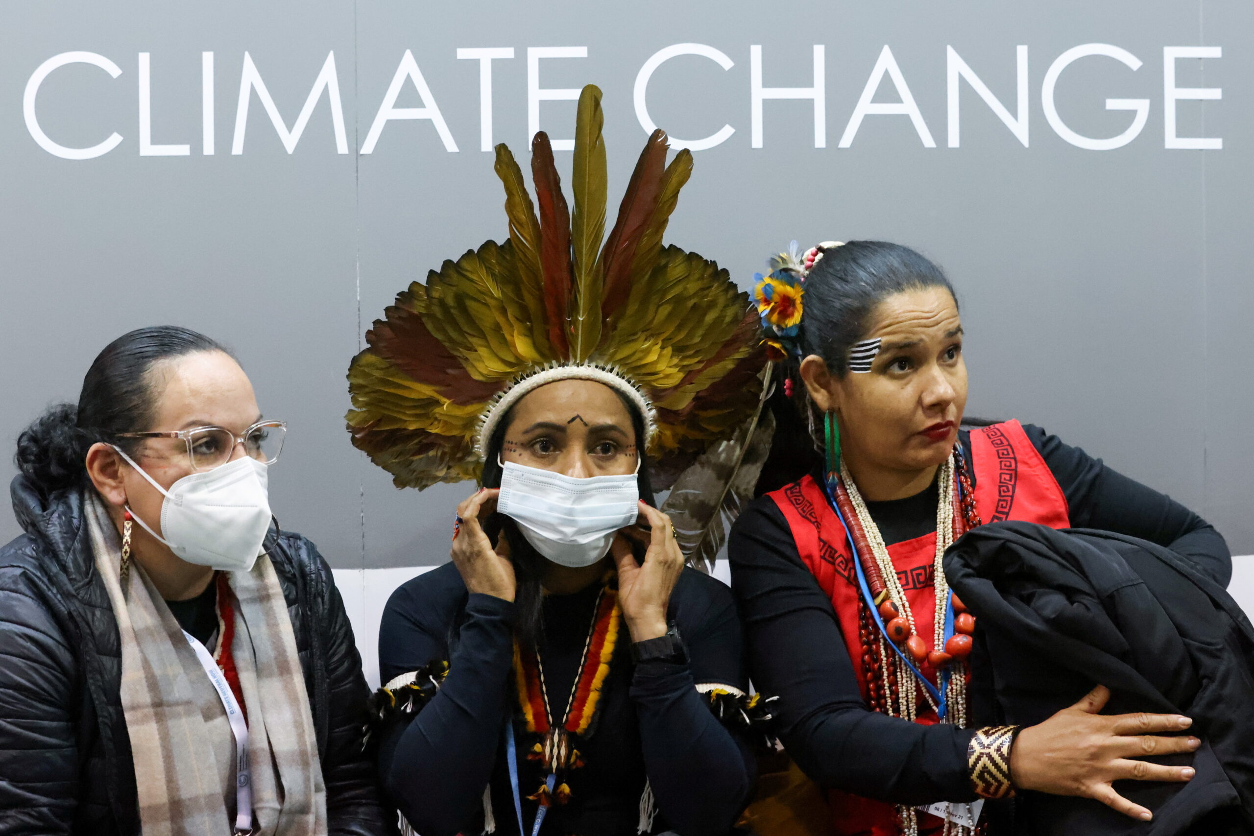 Três mulheres indígenas estão lado a lado. No meio, uma delas usa cocar. Duas estão com máscara contra a covid-19 no rosto. Atrás, há um painel em que se lê, em inglês, ‘mudança climática’.