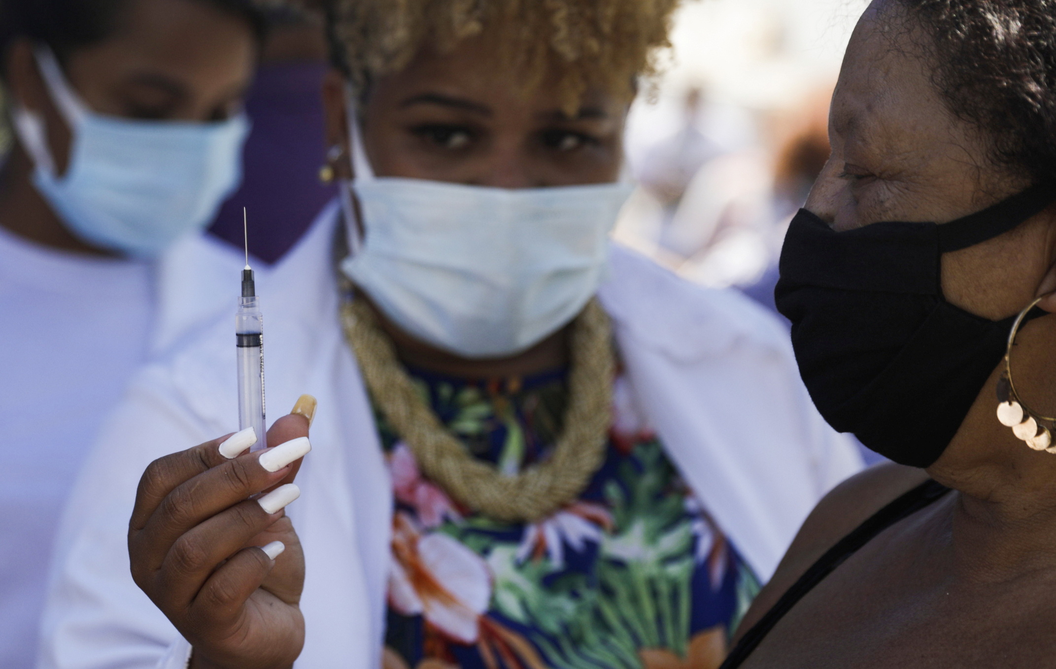 Imagem mostra enfermeira de jaleco branco e máscara segurando seringa com dose da Coronavac e mostrando para idosa antes de vaciná-la