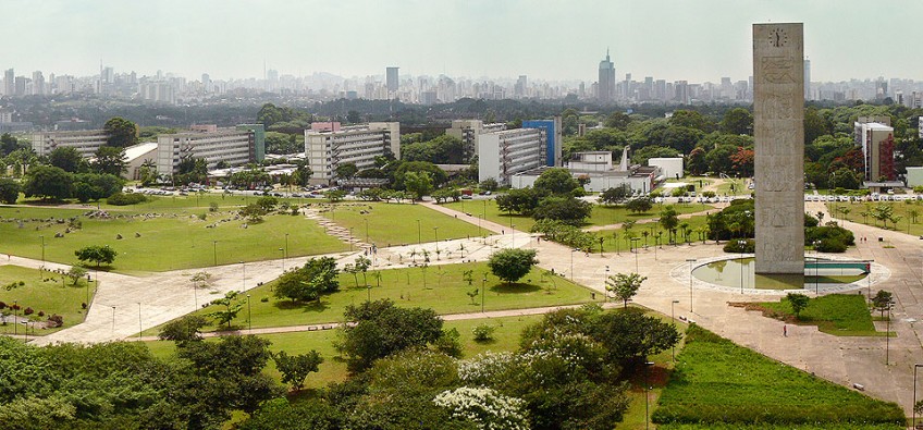 Praça do Relógio tem áreas arborizadas e uma torre com um relógio no centro. Ao fundo, é possível ver alguns arranha-céus da cidade de São Paulo