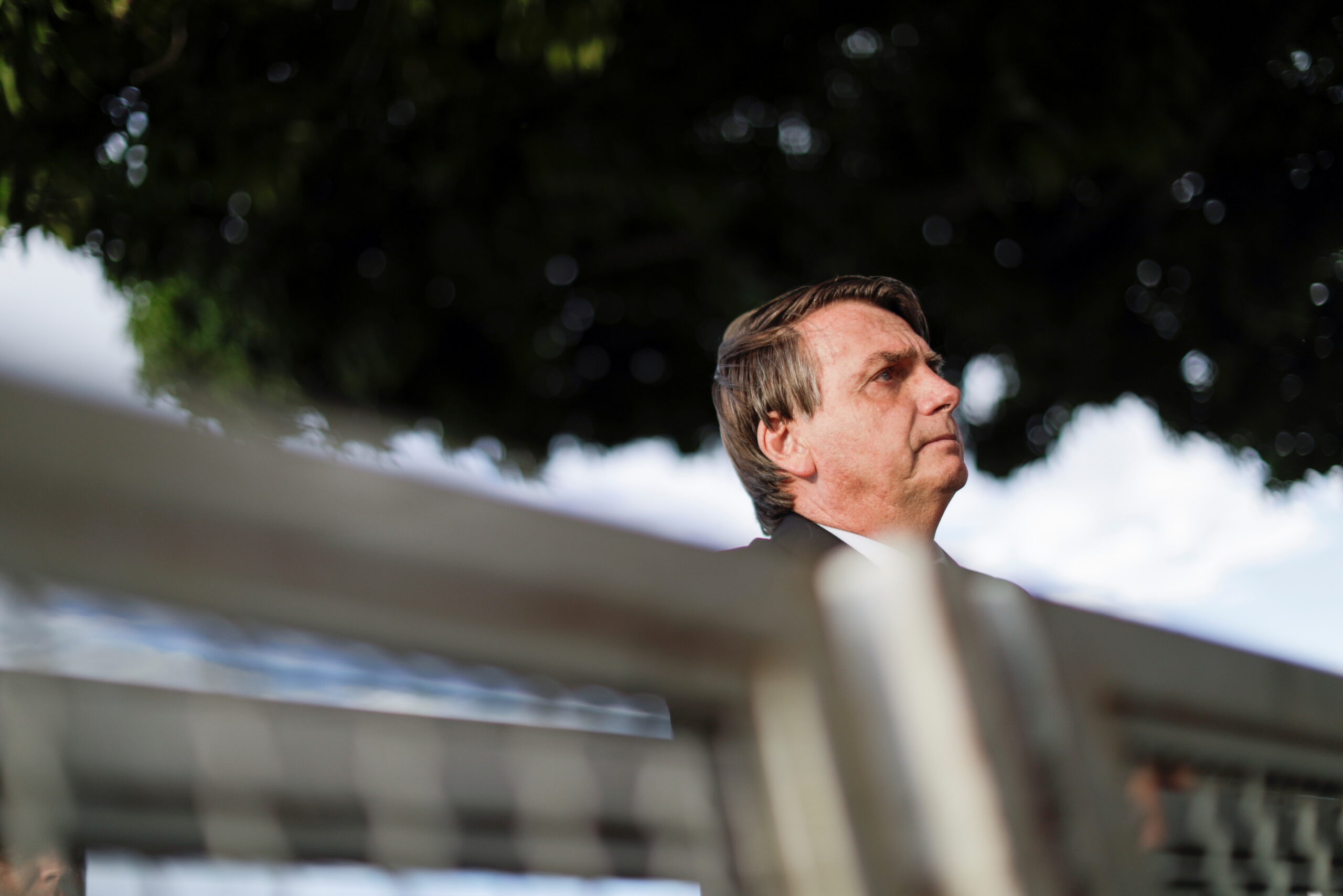 No primeiro plano, há uma grade. A imagem, tirada de baixo, mostra Bolsonaro no segundo plano, com apenas a cabeça sobre a grade