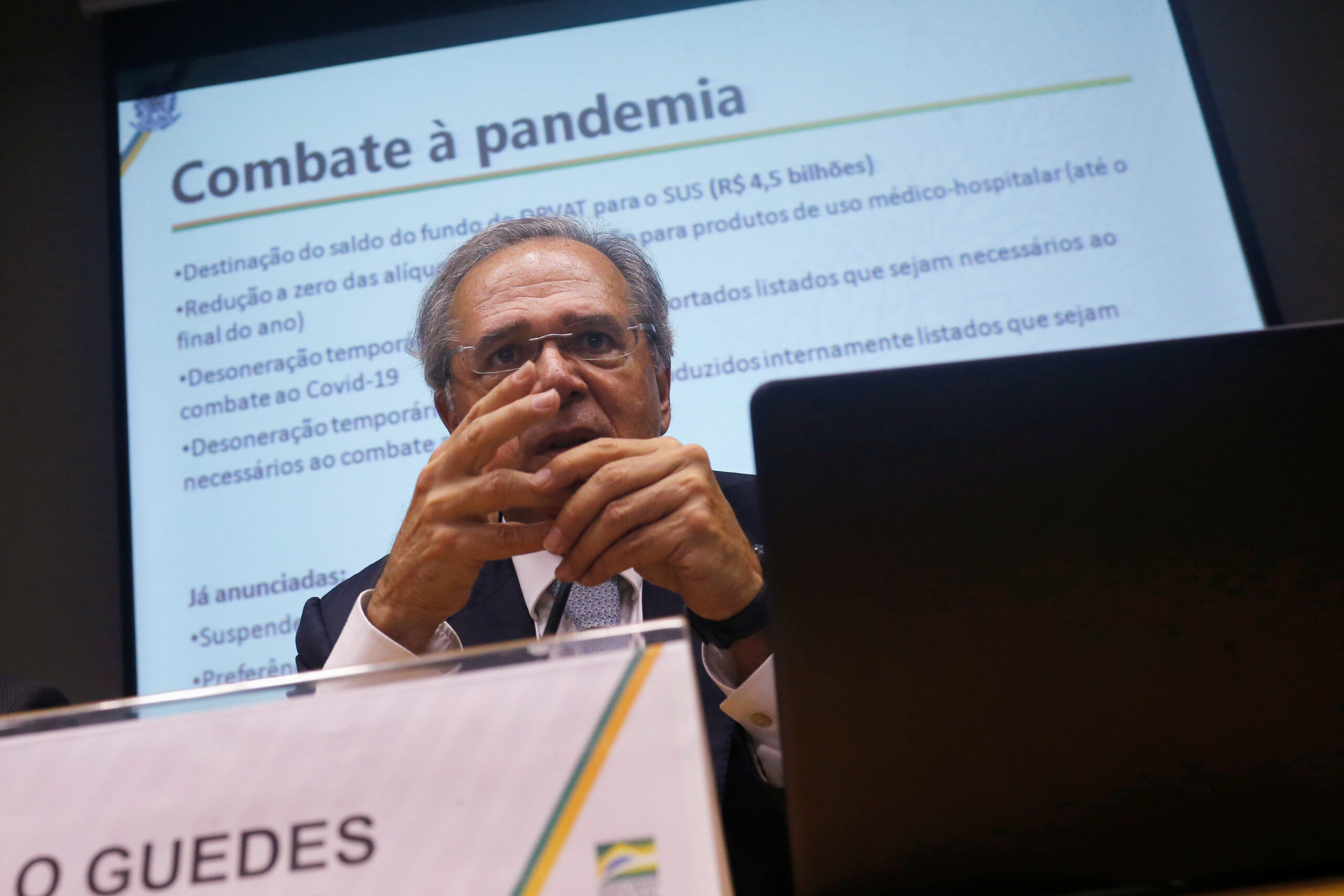 Guedes gesticula atrás de um computador. Ao fundo, tela apresenta slide "combate à pandemia"