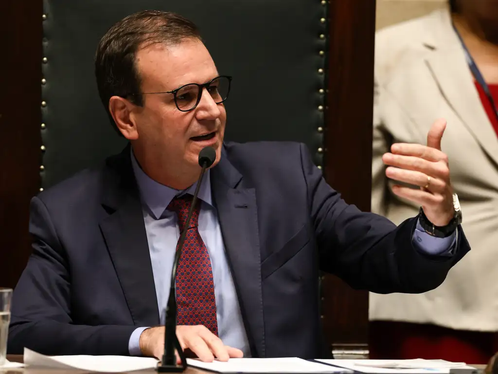 Eduardo Paes (PSD), prefeito do Rio de Janeiro, no dia da sua posse em 1° de janeiro de 2025. Ele está sentado em uma cadeira e fala em um microfone. É um homem branco, usa óculos de grau, terno azul e gravata vermelha