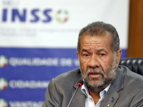O ministro da Previdência Social, Carlos Lupi, em entrevista durante lançamento do cartão Meu INSS Vale+. Ele está sentado, fala em um microfone com um banner escrito INSS ao fundo