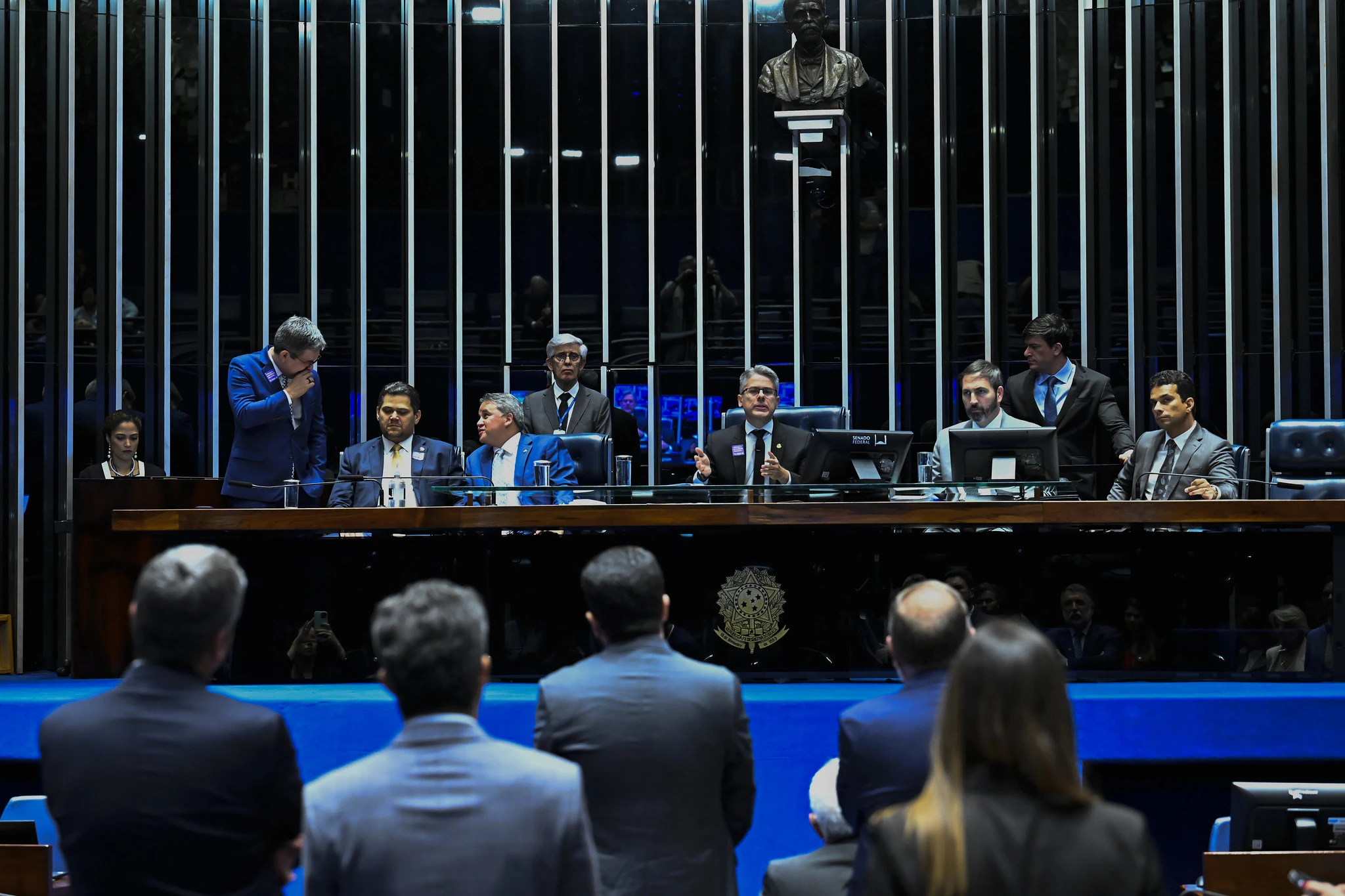 Sessão plenário do Senado, que aprovou o PL da Adultização. Diversas pessoas estão atrás de uma mesa de madeira, sentados. À frente, pessoas em pé e de costas para a câmera