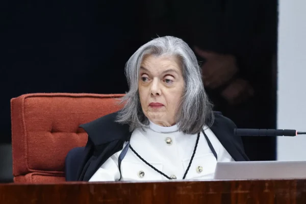 Mulher branca, de cabelos grisalhos e médios, está sentada à mesa. Ela usa toga.