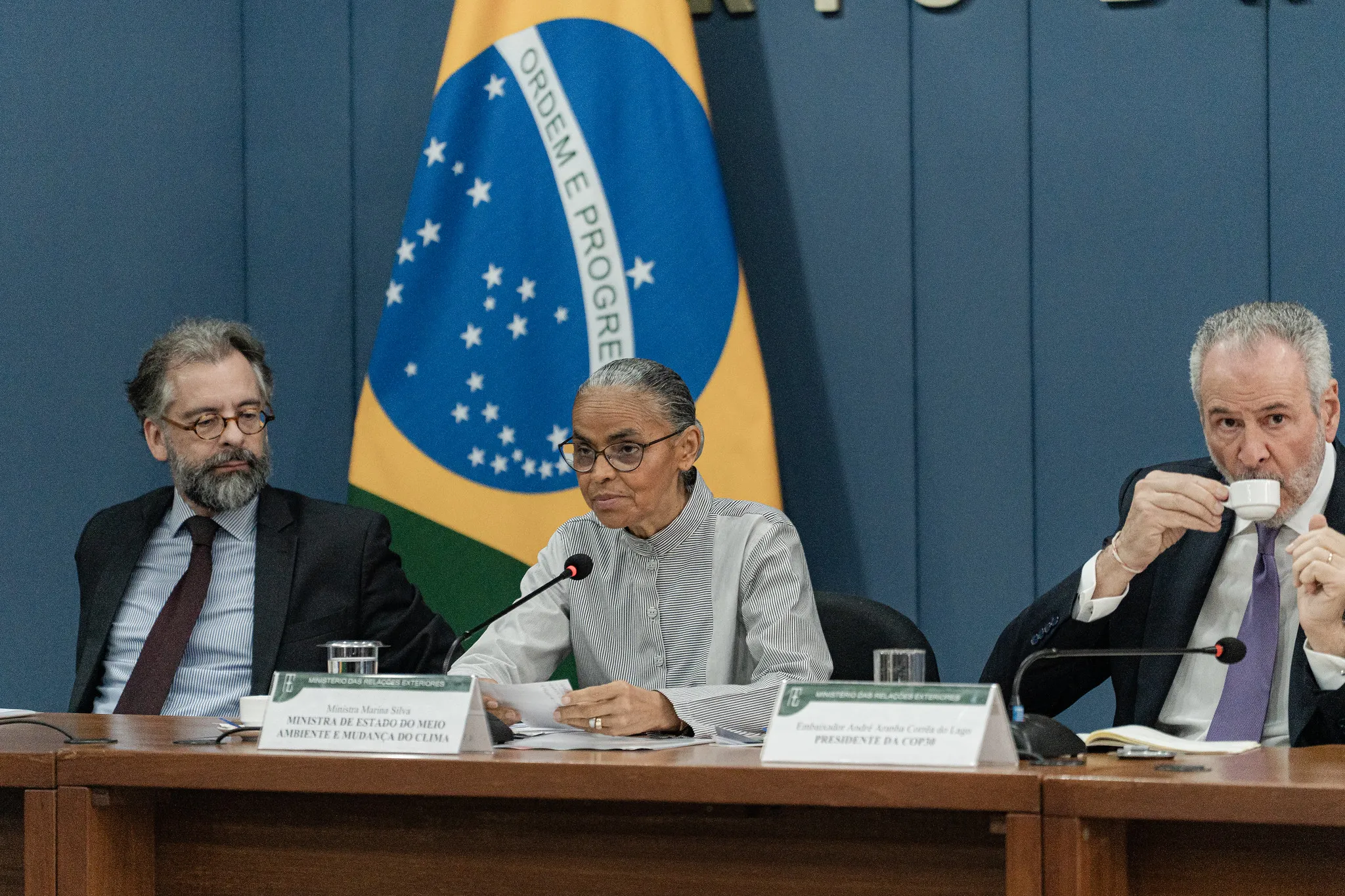 Três pessoas, dois homens e uma mulher, sentam-se lado a lado à mesa e falam ao microfone. Atrás deles, está a bandeira do Brasil.