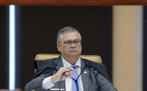 Flávio Dino em sessão plenária do STF. Ele é um homem pardo, de cabelos grisalhos, usa óculos de grau, terno e gravata e uma capa preta. Está sentado e segura microfone com uma das mãos