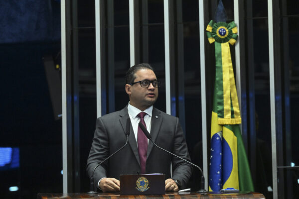 O senador Weverton Rocha (PDT-MA) em discurso na tribuna do Senado. Ele é um homem branco, de cabelos pretos, usa terno e gravata, óculos de grau e fala em um microfone. Ao fundo, uma bandeira do Brasil verde e amarela
