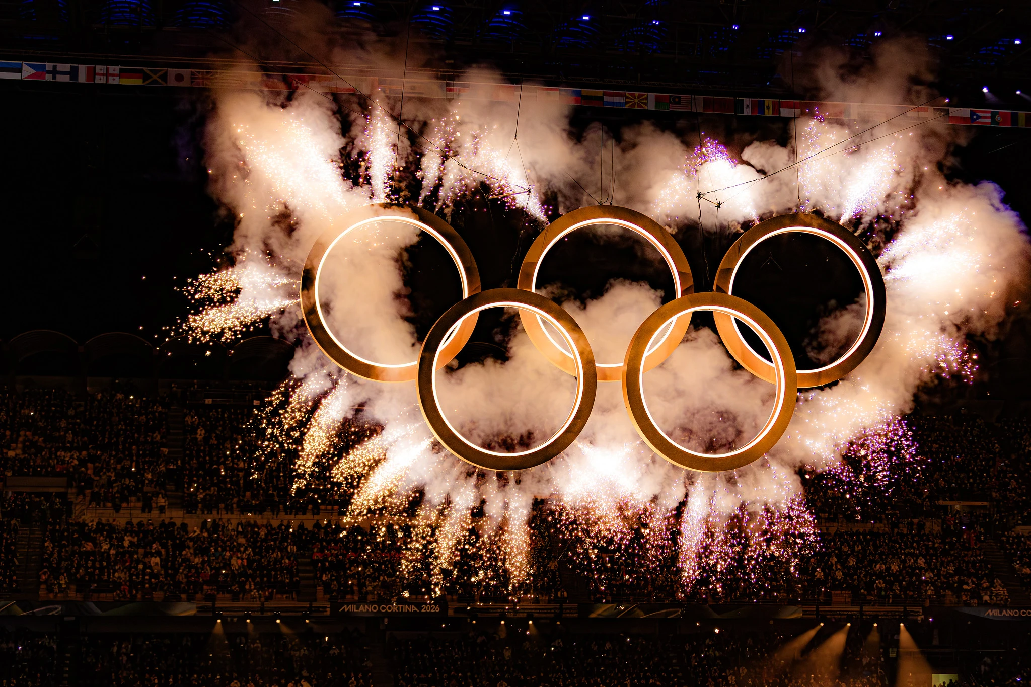Anéis olímpicos na cerimônia de abertura dos Jogos Olímpicos de Inverno de Milão-Cortina. Cinco arcos estão com fogos de artificio ao redor de cada um