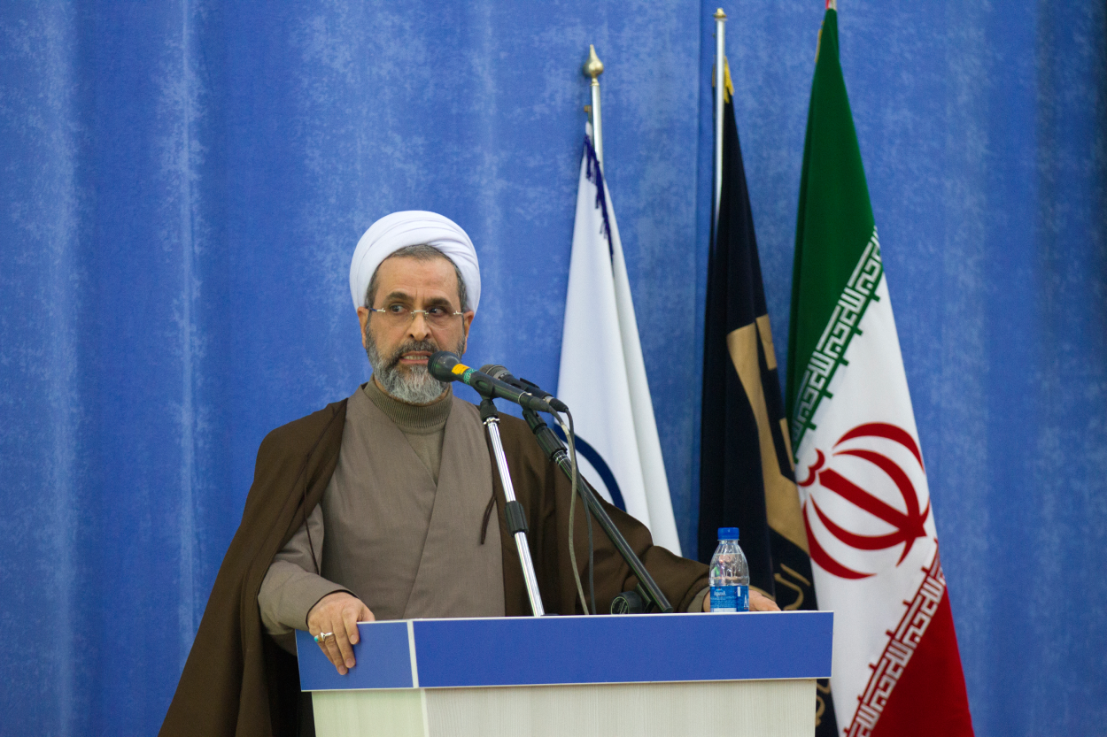 O aiatolá Alireza Arafi. Um homem de turbante está em frente a três bandeiras. Ele está atrás de um púlpito