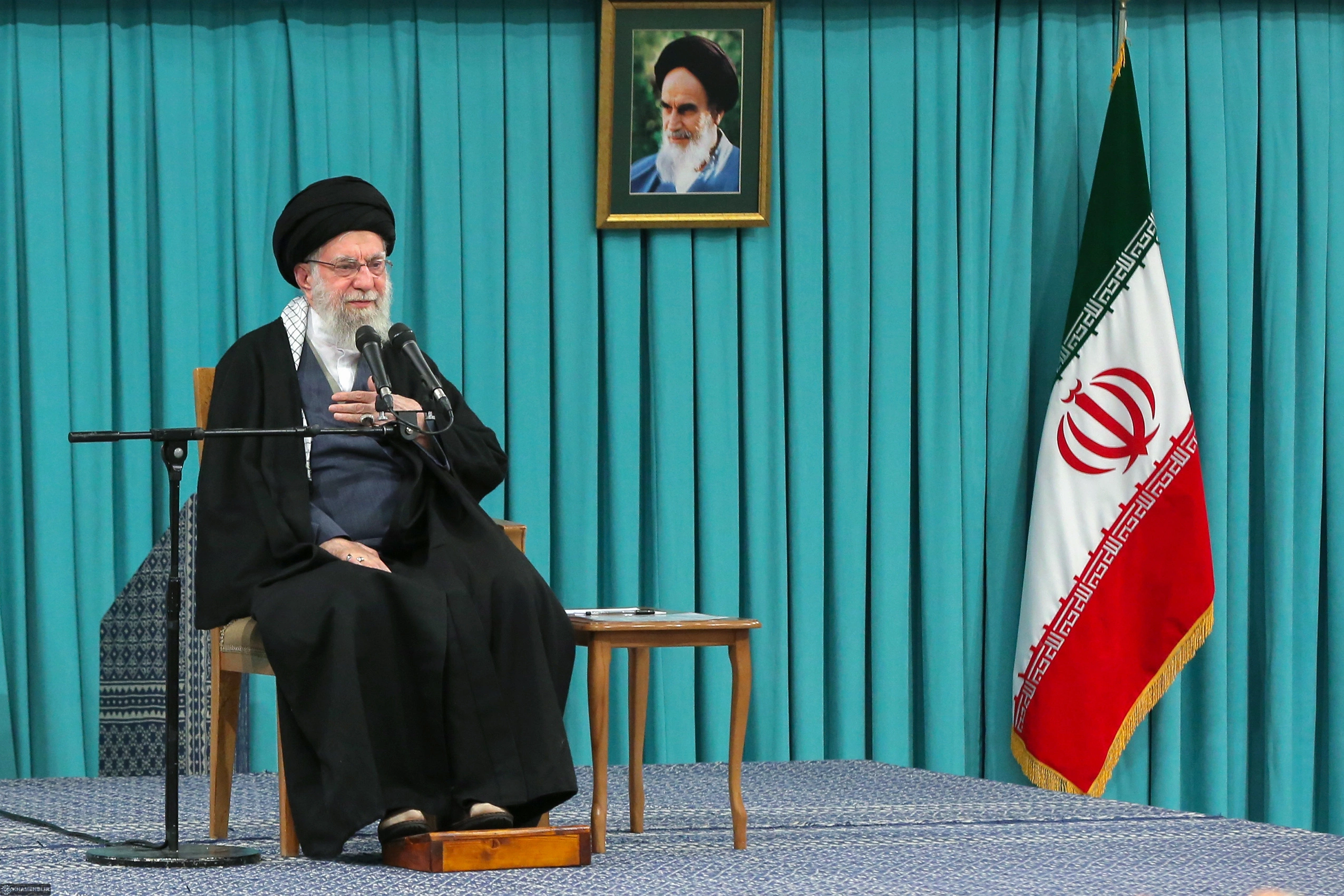 Ali Khamenei, ex-líder supremo do Irã. Um homem está sentado em uma cadeira. Ao fundo, um painel azul e uma bandeira verde, branca e vermelha
