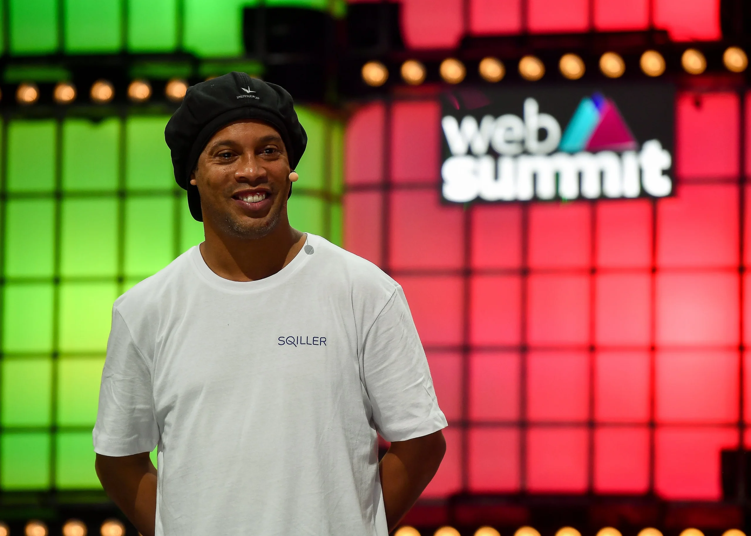 Ronaldinho Gaúcho no Web Summit 2019, em Lisboa, Portugal. Um homem negro está com camiseta branca e uma touca. Ao fundo, um painel vermelho e verde