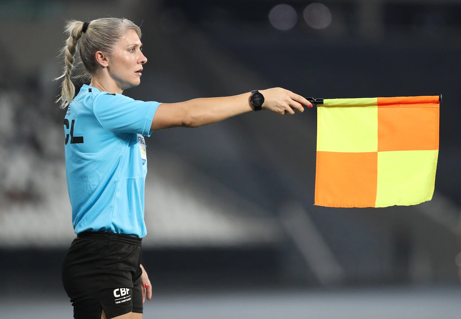 A árbitra assistente Neuza Inês Back. Uma mulher de camisa azul e shorts preto segurando uma bandeira amarela e laranja