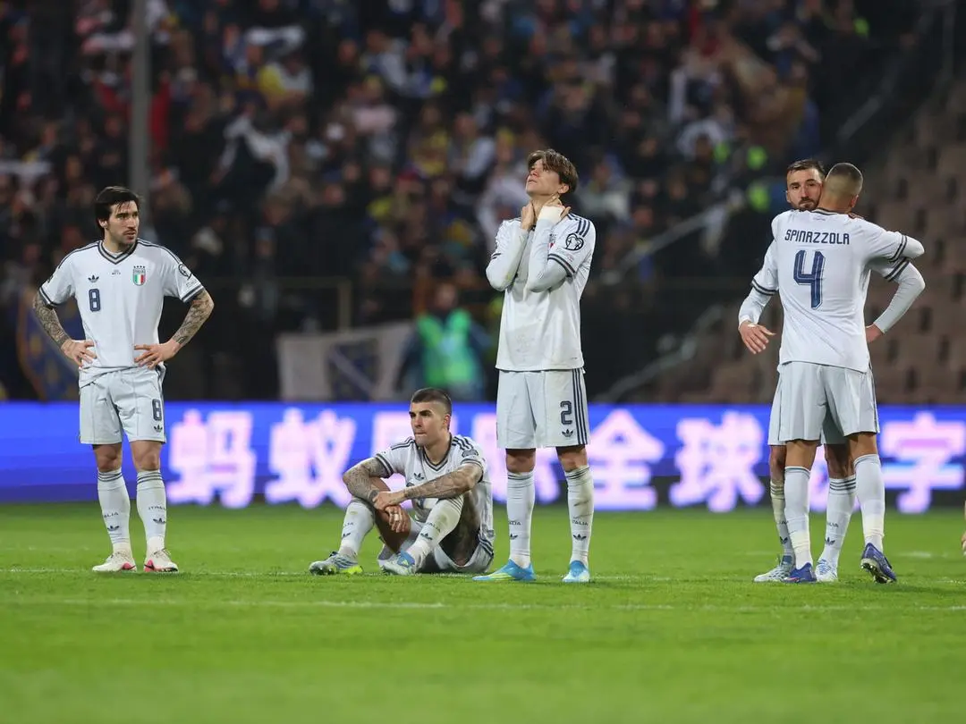 Jogadores da Itália após a disputa por pênaltis contra a Bósnia, pela repescagem europeia da Copa do Mundo de 2026. Homens de uniforme branco chorando. Dois se abraçam. Eles estão em um gramado