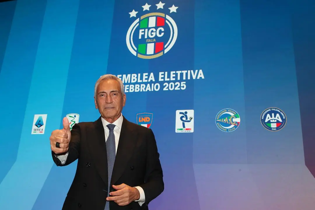 O presidente da FIGC, Gabriele Gravini. Um homem idoso, de terno, faz um jóia com a mão. Ao fundo, um painel azul