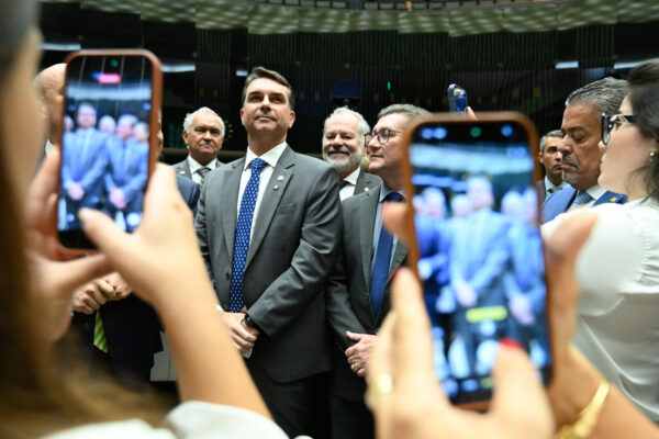 Flávio Bolsonaro (PL-RJ) durante sessão conjunta do Congresso Nacional. Ele é um homem branco, de cabelos pretos, usa terno cinza e gravata azul. Está cercado por pessoas que tiram fotos dele no celular