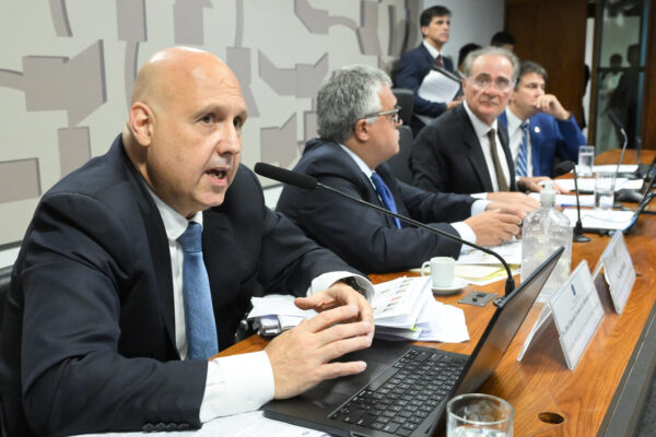 Otto Lobo e senadores em sabatina para a presidência da CVM. Ele é um homem branco, careca, usa terno e gravata, está sentado e fala em um microfone com outros senadores sentados ao lado